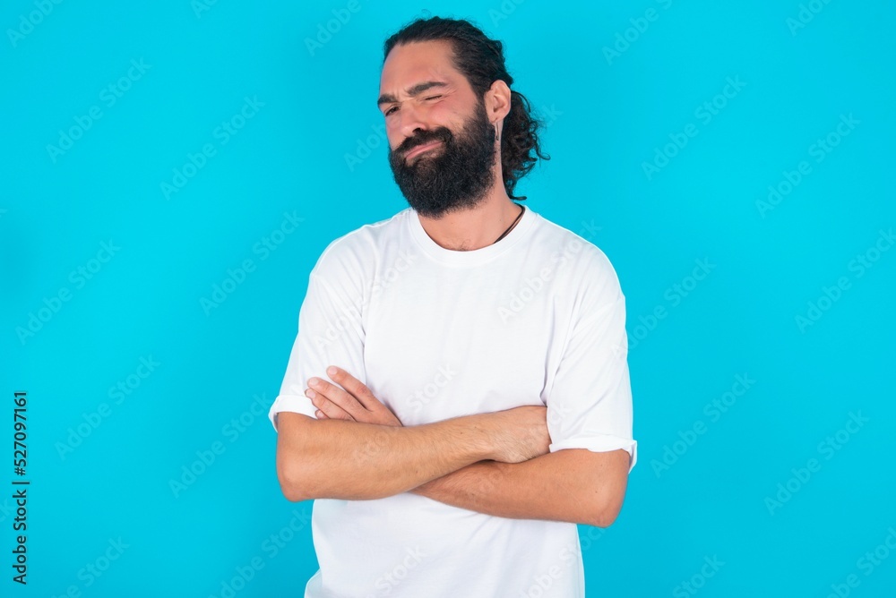 Picture of angry young bearded man wearing white T-shirt over blue studio background crossing arms. Looking at camera with disappointed expression. Picture of angry young bearded man wearing white T-shirt over blue studio background crossing arms. Looking at camera with disappointed expression.