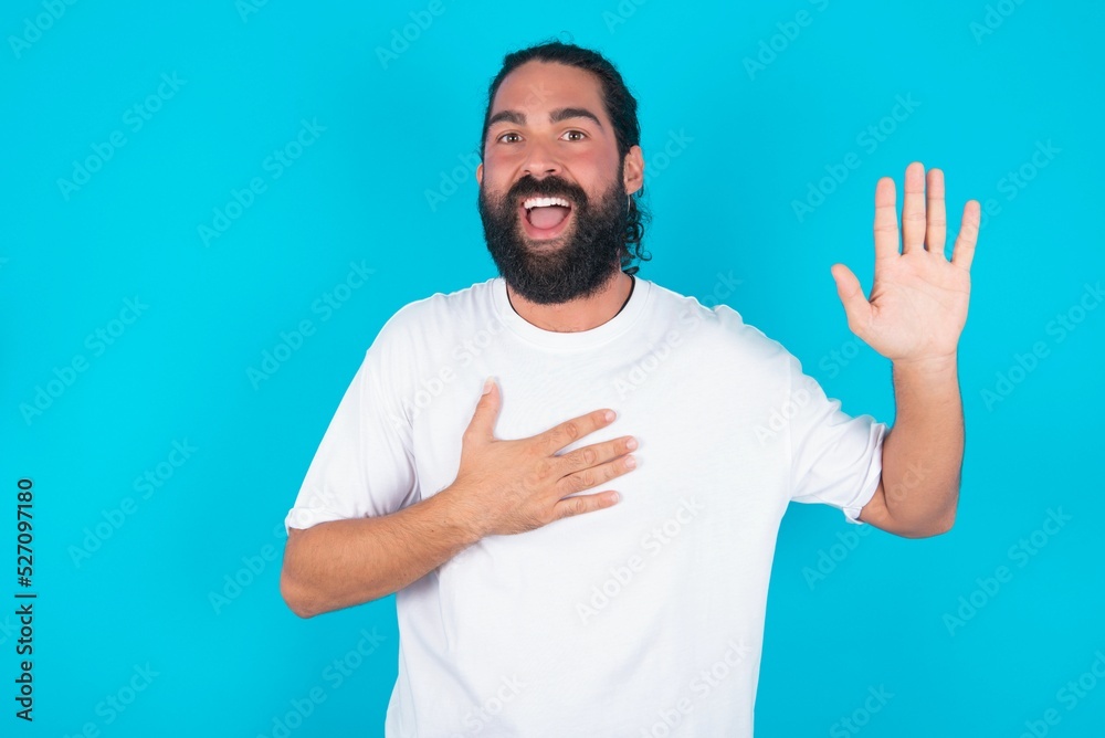 I swear, promise you not regret. Portrait of sincere young bearded man wearing white T-shirt over blue studio raising one arm and hold hand on heart as give oath, telling truth, want you to believe.