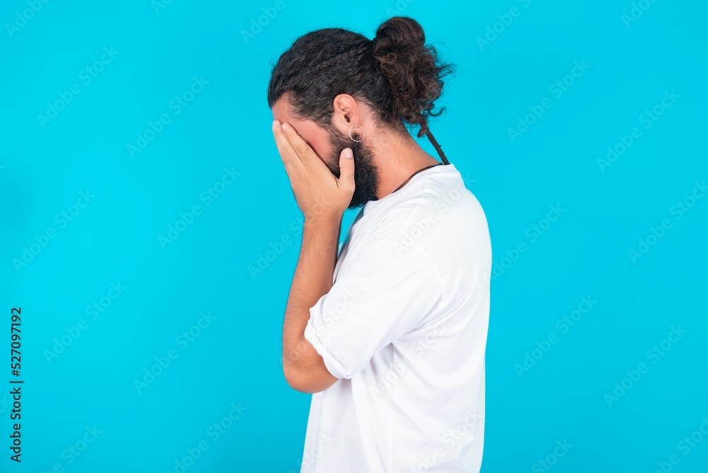 Sad young bearded man wearing white T-shirt over blue studio background covering face with hands and crying. Sad young bearded man wearing white T-shirt over blue studio background covering face with hands and crying.