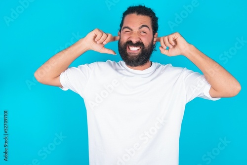 Happy young bearded man wearing white T-shirt over blue studio background ignores loud music and plugs ears with fingers asks to turn off sound