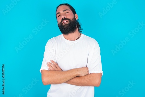 Canvas Print young bearded man wearing white T-shirt over blue studio background frowning his face in displeasure, keeping arms folded, waiting for an explanation