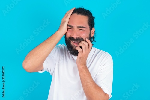 Canvas Print Young gloomy young bearded man wearing white T-shirt over blue studio background, hiding face with hands pouting and crying, standing upset and depressed complaining about job problem