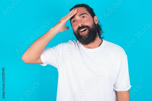 young bearded man wearing white T-shirt over blue studio background touching forehead, hears something surprising, glad receive good news, feels relieved. Almost got in trouble.