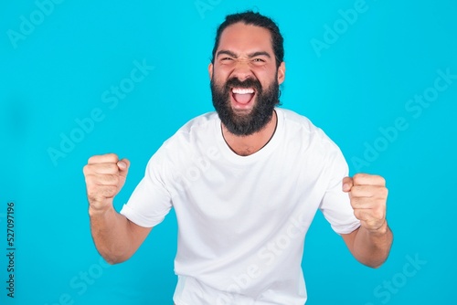 Portrait of young bearded man wearing white T-shirt over blue studio background looks with excitement at camera, keeps hands raised over head, notices something unexpected reacts on sudden news.