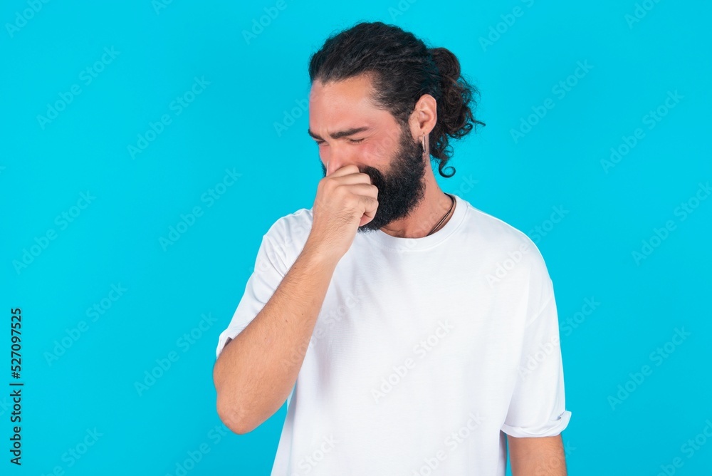 young bearded man wearing white T-shirt over blue studio background, holding his nose because of a bad smell. young bearded man wearing white T-shirt over blue studio background, holding his nose because of a bad smell.