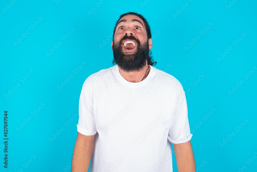 Surprised young bearded man wearing white T-shirt over blue studio background, shrugs shoulders, looking sideways, being happy and excited. Sudden reactions concept. Surprised young bearded man wearing white T-shirt over blue studio background, shrugs shoulders, looking sideways, being happy and excited. Sudden reactions concept.