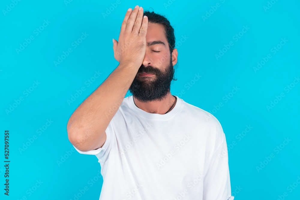 Frustrated young bearded man wearing white T-shirt over blue studio background holding hand on forehead being depressed regretting what he did having headache, looking stressful. Frustrated young bearded man wearing white T-shirt over blue studio background holding hand on forehead being depressed regretting what he did having headache, looking stressful.