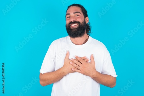 Honest young bearded man wearing white T-shirt over blue studio background keeps hands on chest, touched by compliment or makes promise, looks at camera with great pleasure.