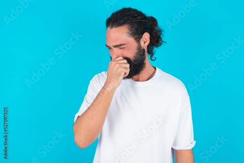 young bearded man wearing white T-shirt over blue studio background  holding his nose because of a bad smell.