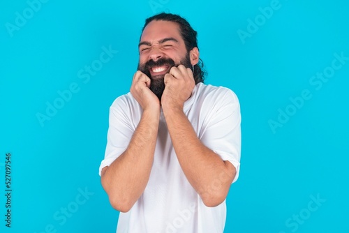 Portrait of young bearded man wearing white T-shirt over blue studio background being overwhelmed, expressing excitement and happiness with closed eyes and hands near face.