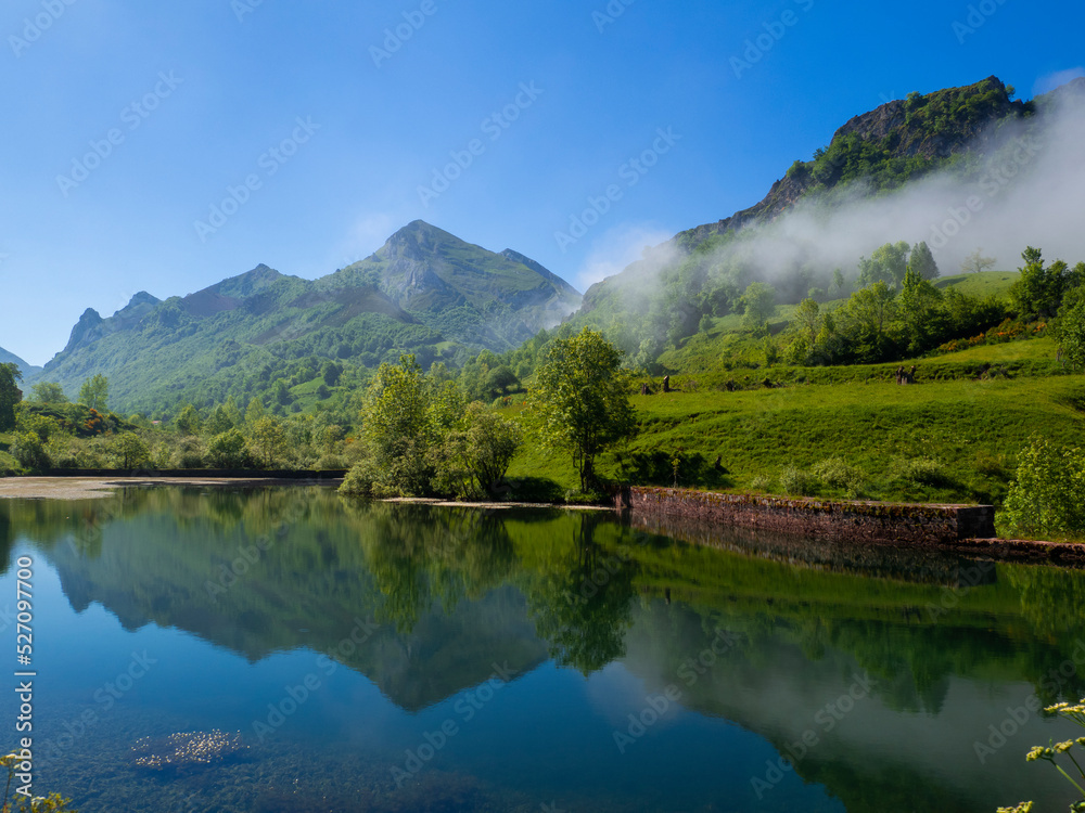 Vistas panorámicas de un paisaje con un lago, reflejos de las montañas y el cielo azul, con niebla blanca y árboles verdes en Asturias, España, verano de 2021.