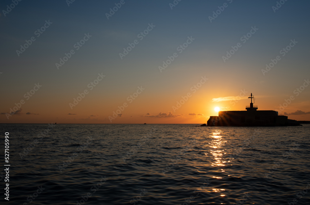 The sun goes down at the sea. The sky glows blue and orange. The light is reflected on the waves. The shadow of a signal tower is in front of the sun at the side of the picture.