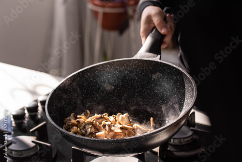 Close up of professional Chef cook hands roasts champignons mushrooms with cream in wok pan for Mediterranean cuisine. 