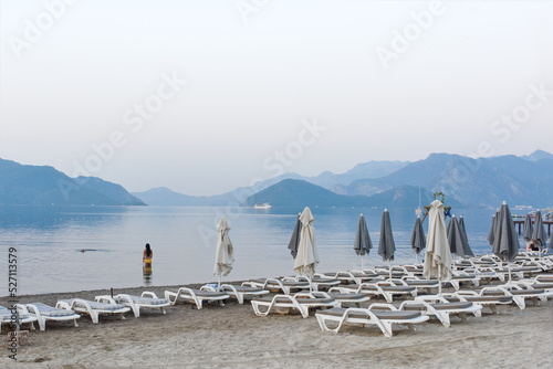 Fototapeta Naklejka Na Ścianę i Meble -  Beach chairs and umbrellas overlooking the bay in the early morning. Calm on the sea in the bay of Marmaris. Morning swim in the sea. Turkey, summer, beach vacation.