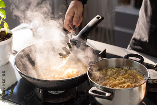 Professional chef cook making Italian Tagliatelle pasta with mushrooms and cream at modern kitchen gas stove in wok pan and pot.