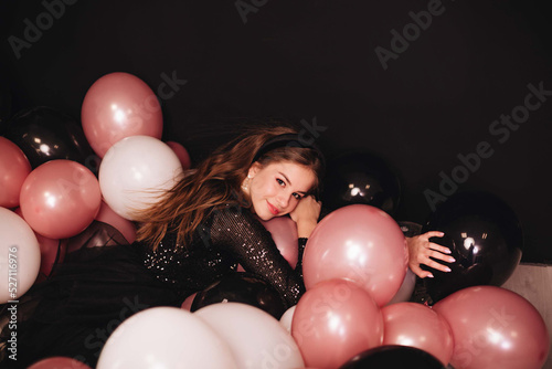 A girl with long hair in a black shiny dress celebrates her birthday with a lot of colorful balloons isolated on a black background. Copy space.
