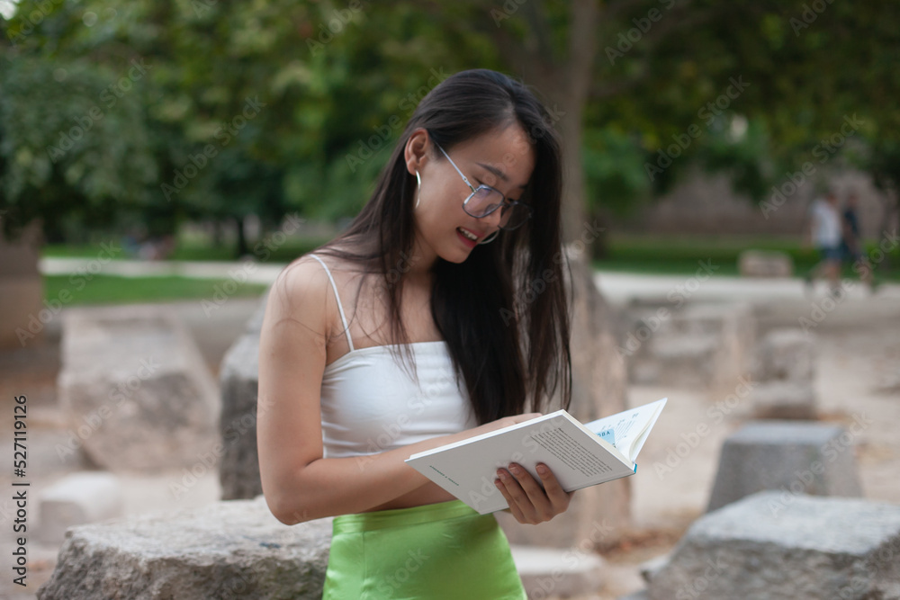Fototapeta premium Beautiful young asian girl wearing glasses studying arts at college or university working on tasks in the green park or campus. Young Chinese student writing and drawing. Female education in Asia.