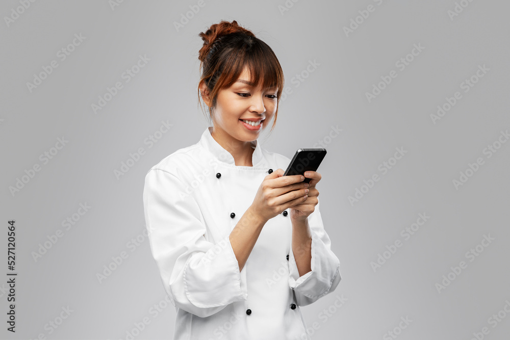 cooking, culinary and people concept - happy smiling female chef in white jacket with smartphone over grey background