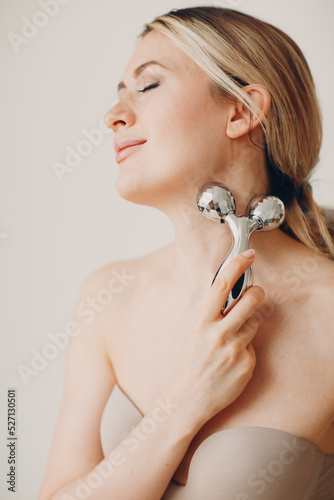 Beautiful young adult woman with massage face roller on beige background
