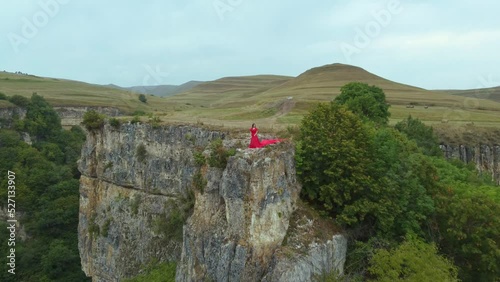 A girl in a red dress stands on the edge of a cliff, summer, mountains and forest, a seed from a drone