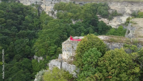 A girl in a red dress stands on the edge of a cliff, summer, mountains and forest, a seed from a drone