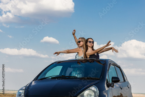 Beautiful young women in sunglasses dance against the background of a blue sky with clouds leaning out of the car hatch. The concept of traveling by private car