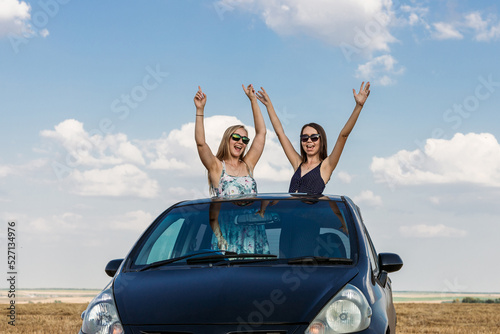Beautiful brunette and blonde in dark glasses are traveling by car and enjoying the ride.Young women have fun dancing out of the car hatch