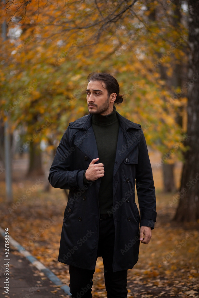 Handsome long haired man in black trench coat walking in the park in wonderful autumn day. Fashion and style concept