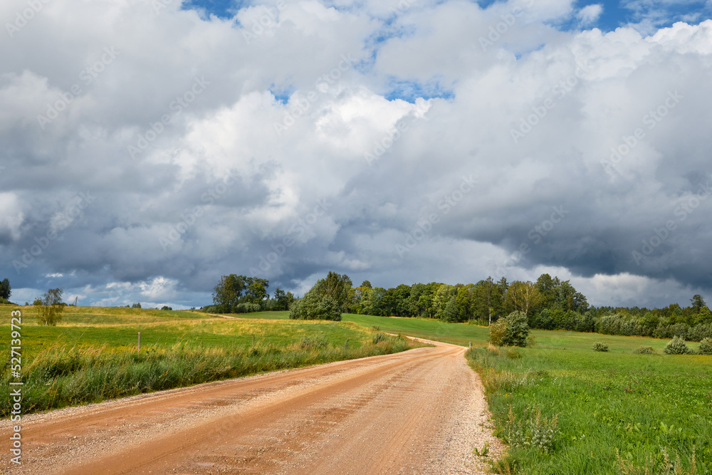 Gravel road, dirt track through Estonian countryside in Summer. Green fields and forest under dramatic sky with clouds. Gravel road, dirt track through Estonian countryside in Summer. Green fields and forest under dramatic sky with clouds.