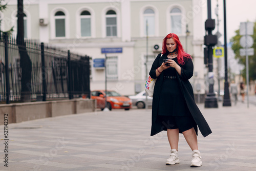 European plus size woman walking outdoor street city with mobile cellular phone. Young red pink haired body positive girl using cellphone smartphone