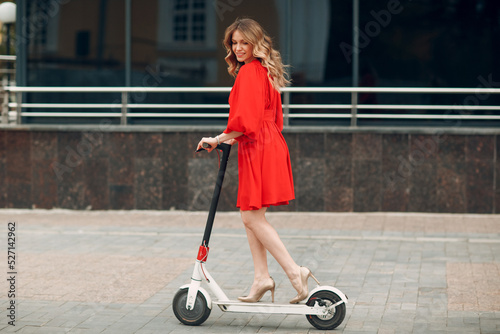 Young woman riding electric scooter in red dress at the city.