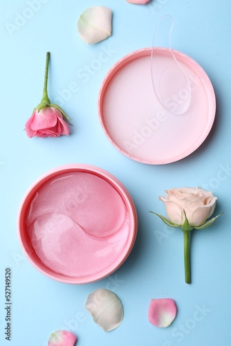 Under eye patches in jar and rose flowers on light blue background, flat lay. Cosmetic product