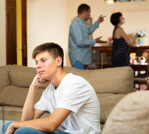 Portrait of upset tired teen boy sitting at home on background of his arguing parents