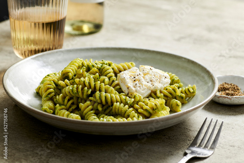 Bowl of pesto pasta topped with creamy burrata next to a glass o photo