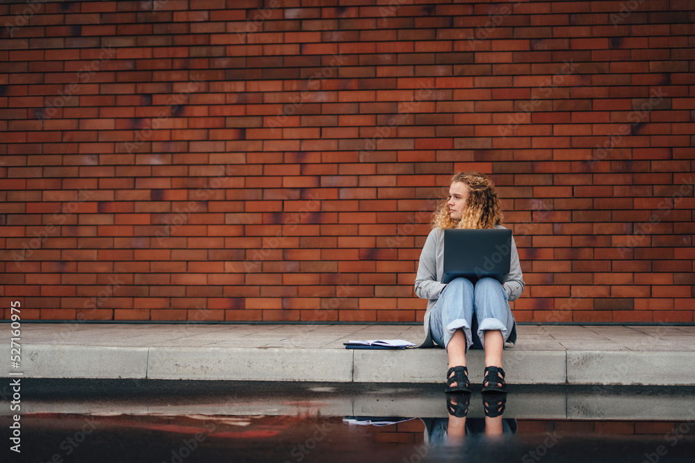 Caucasian girl having training course via notebook device, sitting outdoors in city street on the background of a brick wall with free space for text Caucasian girl having training course via notebook device, sitting outdoors in city street on the background of a brick wall with free space for text