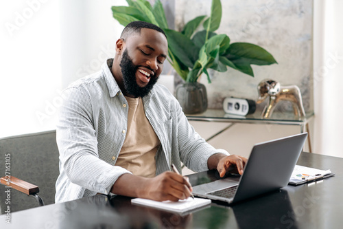 Happy joyful successful clever african american business man, sales manager, sitting at a desk in modern office, working on a project, analyzing profit, taking notes, prepares a report, smiling