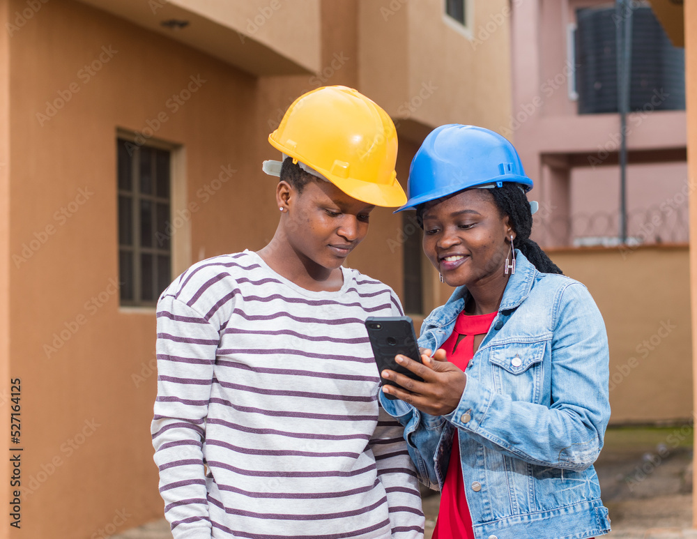 African ladies, women or coworkers from Nigeria with safety helmet, holding and using a phone as they represents industrial professionals like construction specialist, builders, architect and engineer African ladies, women or coworkers from Nigeria with safety helmet, holding and using a phone as they represents industrial professionals like construction specialist, builders, architect and engineer