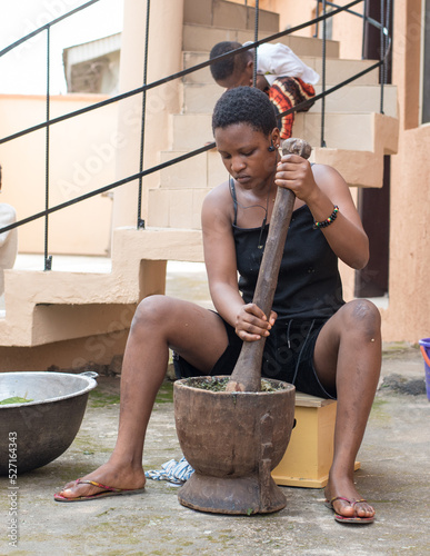 An African young girl, lady or woman from Nigeria using a wooden pestle and mortar to pound and grind edible vegetables for food cooking, herb or medications