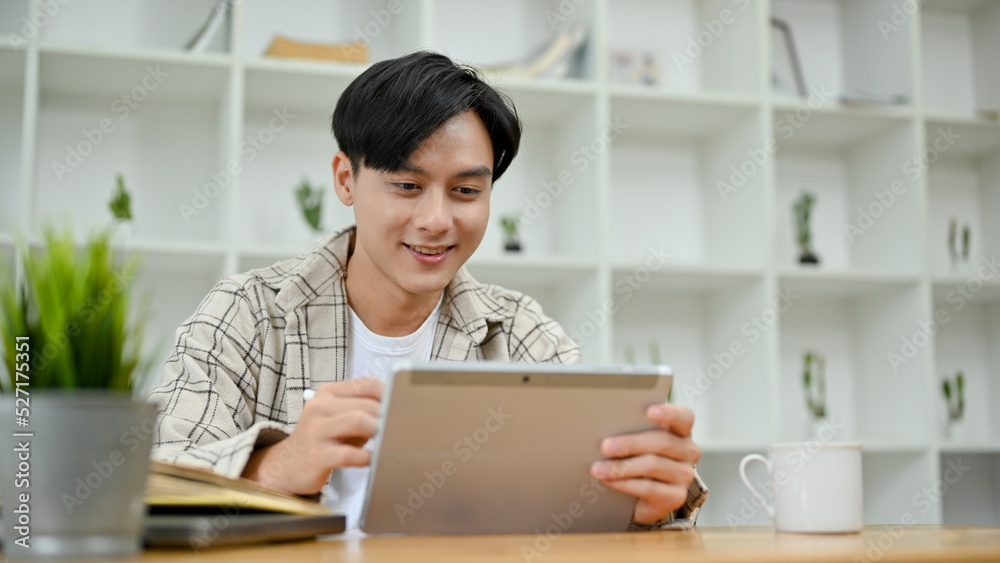 Handsome Asian man in causal outfit using digital tablet touchpad at his desk in office room. Handsome Asian man in causal outfit using digital tablet touchpad at his desk in office room.