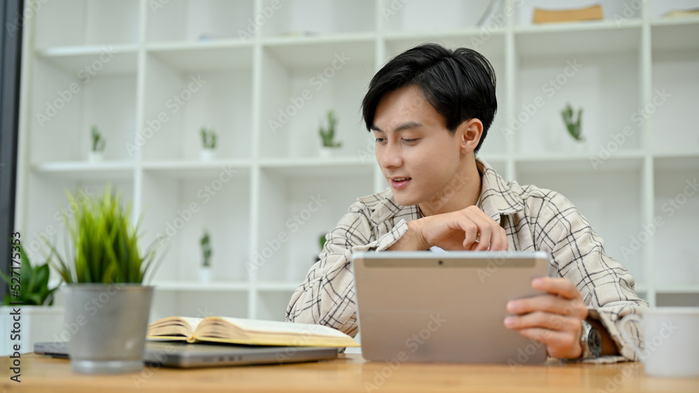 Young Asian male freelancer using digital tablet touchpad while reading book. Young Asian male freelancer using digital tablet touchpad while reading book.