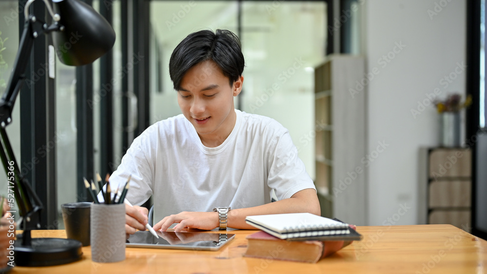 Handsome Asian male university student concentrating doing his homework on tablet Handsome Asian male university student concentrating doing his homework on tablet