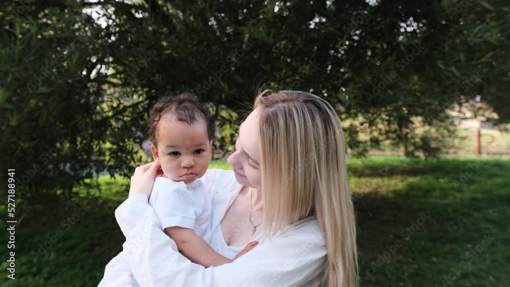 A Caucasian mother gently hugs her African child and holds him in her arms while walking in the park. Mixed multiracial family A Caucasian mother gently hugs her African child and holds him in her arms while walking in the park. Mixed multiracial family
