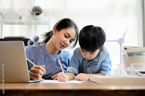 Young Asian mother is an architect spending time with her little kid son practicing pencil drawing and writing on paper at home while multitasking. Mother helping son, Homeschool concept