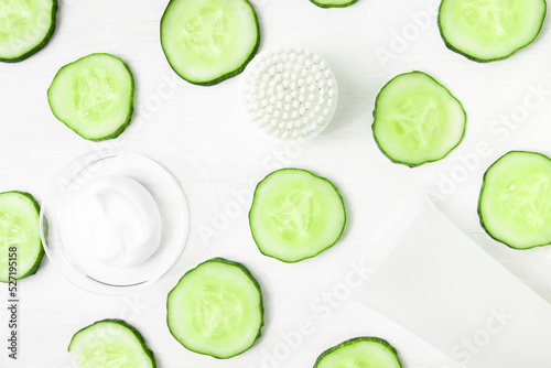 Cosmetic background with cucumber slices and white color cosmetic product and facial cleansing brush, flat lay.