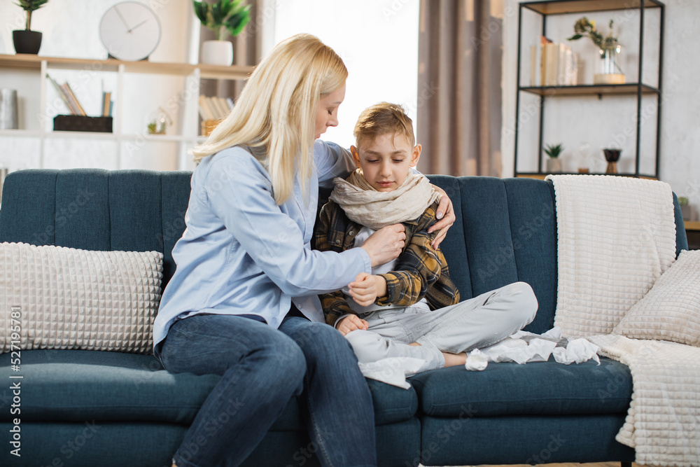Middle aged worried caring blond woman, mother, sitting on sofa at home, hugging her sick sad teen son, measuring temperature using thermometer. Flu, cold, virus infection.