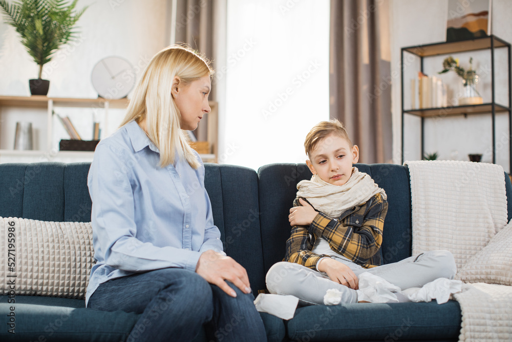 Pleasant 40-aged caucasian mother measuring temperature of her teen boy, which is sitting near her on soft couch with scarf around neck. Flu, covid-19 concept.