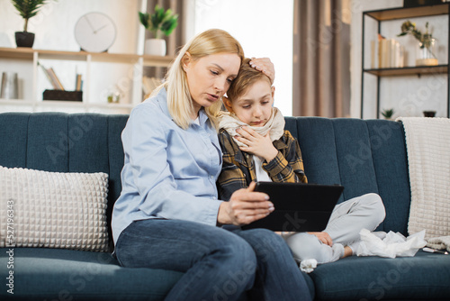 Cute sick teen caucasian boy, putting his head on mother's head, while having online video call with doctor on tablet pc. Flu and cold at home. photo