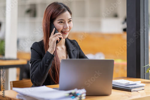 Young Business Asian woman talking on a smartphone and sitting on desk in workplace, Asian female Business accountant Documents data sitting at his workplace.