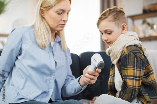Mother taking care of her cute sick teen boy suffering from cold, checking temperature using infrared forehead thermometer gun, looking each other, sitting on the sofa.