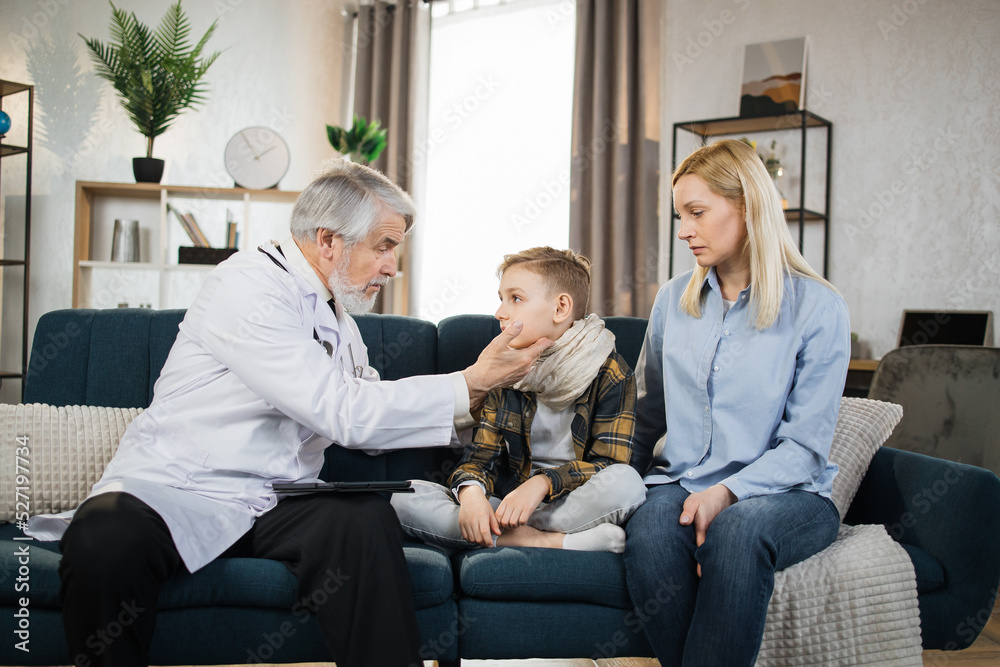 Mature doctor groping checking pain and swelling in the neck sore throat and cough thyroid nodule inflammation of the lymph nodes tonsillitis of sick male patient boy with his mom during home visit Mature doctor groping checking pain and swelling in the neck sore throat and cough thyroid nodule inflammation of the lymph nodes tonsillitis of sick male patient boy with his mom during home visit